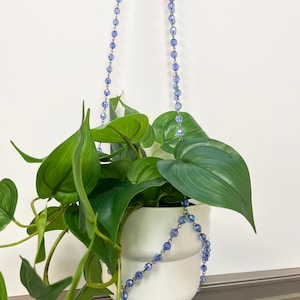 May include: A white ceramic planter with a leafy green plant hanging from a blue beaded chain.