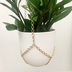 May include: A white ceramic hanging planter with a gold chain. The planter is empty and ready to be filled with a plant.