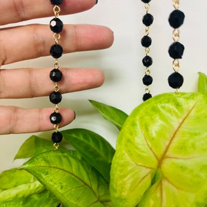 May include: A gold chain necklace with black faceted beads. The necklace is hanging from a plant with large green leaves.