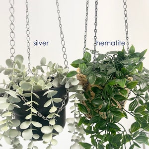 May include: Two hanging planters with different plants. The planter on the left has a silver chain and a black pot with a light green plant. The planter on the right has a hematite chain and a woven basket with a dark green plant.