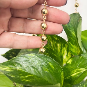 Puede incluir: Un collar de cadena de oro con cuentas redondas. La cadena se sostiene en una mano y cuelga sobre una planta verde.