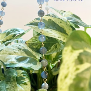 Puede incluir: Primer plano de un collar con cuentas de ágata azul denim ensartadas en una cadena dorada. Las cuentas varían en tonos de azul y gris. El texto "DENIM BLUE AGATE" se muestra en azul.