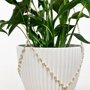 Puede incluir: Una maceta de cerámica blanca con un diseño acanalado está decorada con una cadena dorada enhebrada con perlas blancas. La maceta está llena de una planta verde de hojas.