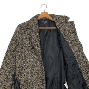 Puede incluir: Un abrigo de tweed en tonos negro, marr&oacute;n y beige cuelga de una percha de madera. El abrigo presenta un cuello con solapas y un forro negro. La marca "nous-rue" es visible en la etiqueta.