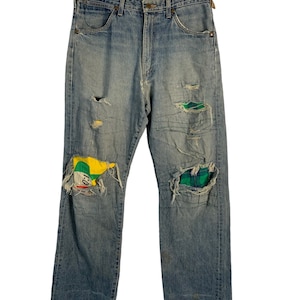 Op de afbeelding: Lichtblauwe spijkerbroek met meerdere patches en scheuren. De patches zijn gemaakt van verschillende stoffen, waaronder ruit en een kleurrijk geometrisch patroon.
