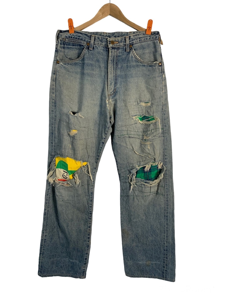 Op de afbeelding: Lichtblauwe spijkerbroek met meerdere patches en scheuren. De patches zijn gemaakt van verschillende stoffen, waaronder ruit en een kleurrijk geometrisch patroon.