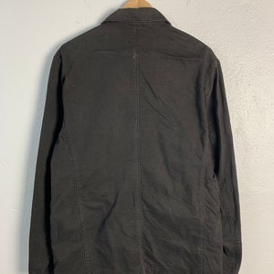 Puede incluir: Chaqueta marr&oacute;n oscuro colgada de una percha de madera. La chaqueta tiene cuello, mangas largas y cierre de botones. La parte trasera muestra costuras verticales y un corte entallado. La chaqueta est&aacute; hecha de una tela resistente.