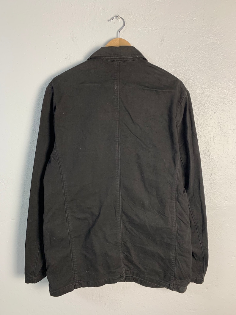 Puede incluir: Chaqueta marr&oacute;n oscuro colgada de una percha de madera. La chaqueta tiene cuello, mangas largas y cierre de botones. La parte trasera muestra costuras verticales y un corte entallado. La chaqueta est&aacute; hecha de una tela resistente.