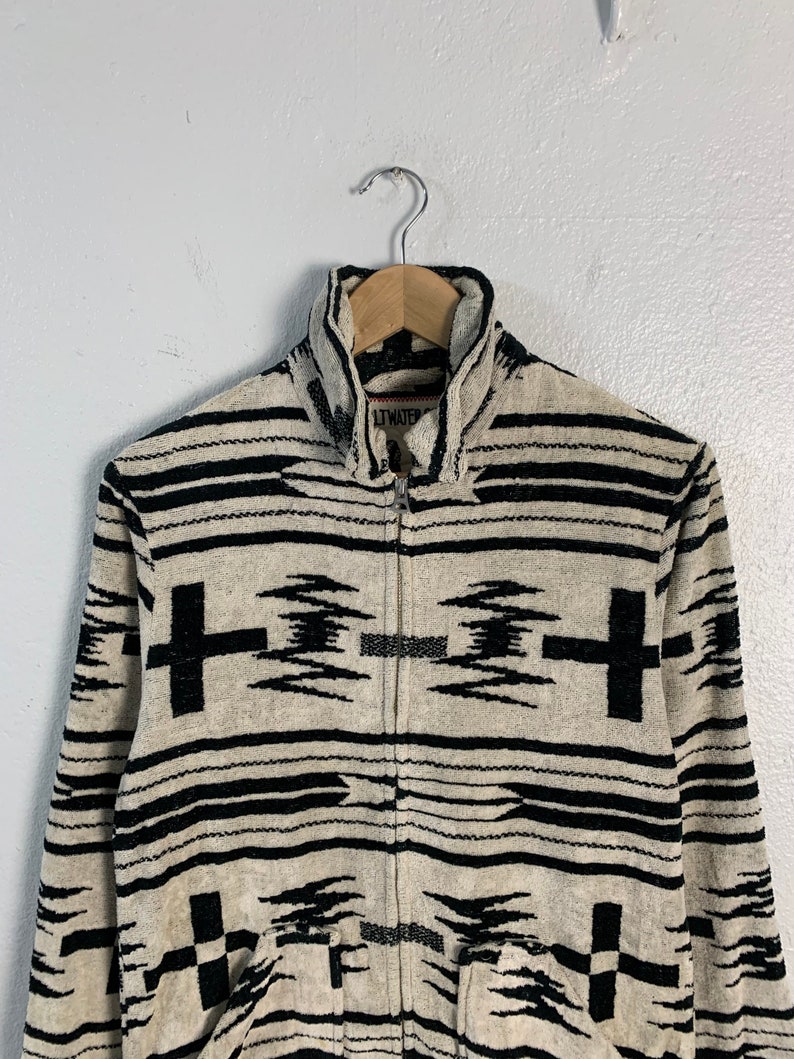 Puede incluir: Una chaqueta con cremallera de color crema con un patr&oacute;n geom&eacute;trico en blanco y negro. La chaqueta tiene un cuello alto y una cremallera. El nombre de la marca "LITWATER" es visible en la parte delantera de la chaqueta.