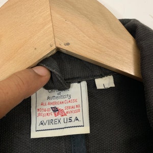 Puede incluir: Primer plano de una chaqueta gris oscuro con una etiqueta que dice "Certificate of Authenticity ALL AMERICAN CLASSIC" y "AVIREX U.S.A.". La etiqueta tambi&eacute;n muestra la bandera estadounidense. La chaqueta est&aacute; colgada en una percha de madera.