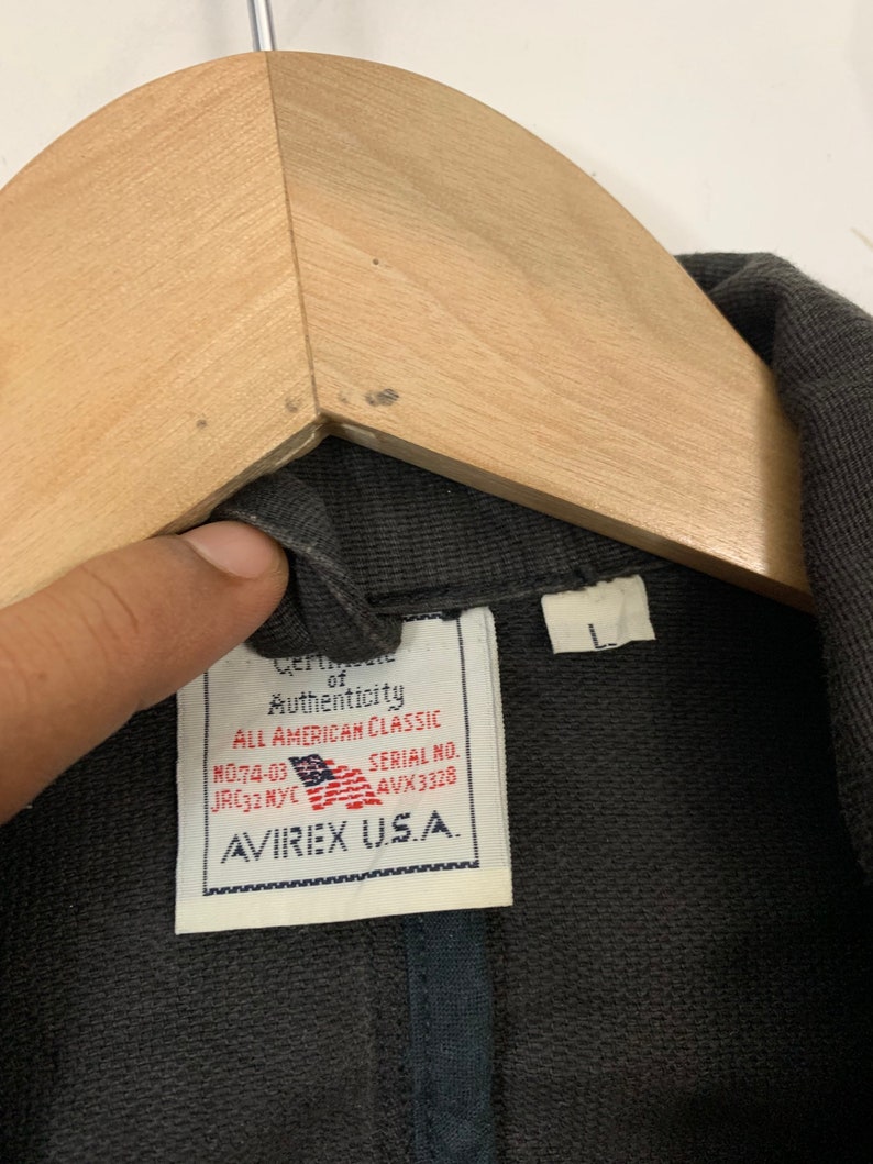 Puede incluir: Primer plano de una chaqueta gris oscuro con una etiqueta que dice "Certificate of Authenticity ALL AMERICAN CLASSIC" y "AVIREX U.S.A.". La etiqueta tambi&eacute;n muestra la bandera estadounidense. La chaqueta est&aacute; colgada en una percha de madera.