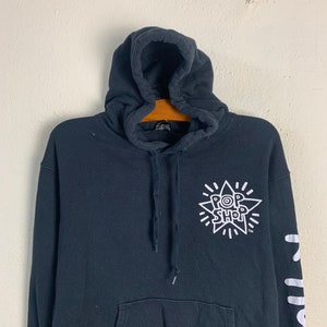 May include: Black hooded sweatshirt with a white starburst graphic featuring the text "Pop Shop" and a trademark symbol. The left sleeve has a white graphic with the text "Pop Shop" repeated vertically.