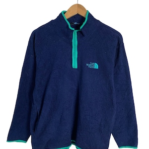 May include: A navy blue fleece pullover with a green zipper and trim. The North Face logo is embroidered on the front in white and blue.