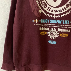 Puede incluir: Una sudadera color granate con un estampado gr&aacute;fico de un s&iacute;mbolo yin yang con la palabra "Hawaii" y el texto "Enjoy Surfin' Life". El gr&aacute;fico tambi&eacute;n incluye el texto "Haleiwa" y "Waimea" con el a&ntilde;o "1971" impreso debajo.