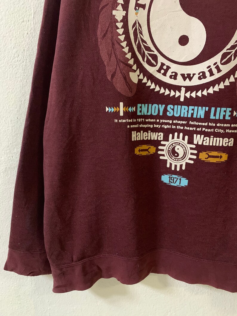 Puede incluir: Una sudadera color granate con un estampado gr&aacute;fico de un s&iacute;mbolo yin yang con la palabra "Hawaii" y el texto "Enjoy Surfin' Life". El gr&aacute;fico tambi&eacute;n incluye el texto "Haleiwa" y "Waimea" con el a&ntilde;o "1971" impreso debajo.