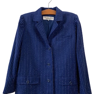 Puede incluir: Una chaqueta azul marino con rayas blancas finas y una etiqueta que dice "Christian Dior" en blanco. La chaqueta tiene un cierre de botón único y dos bolsillos de solapa.