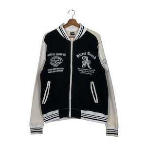 May include: Black and white baseball jacket with a zipper closure. The jacket has white sleeves and a black body with white stripes on the sleeves and bottom. The jacket has a white embroidered logo on the left side of the chest that says "Street Board" and a white embroidered logo on the right side of the chest that says "Seen It Lived It."