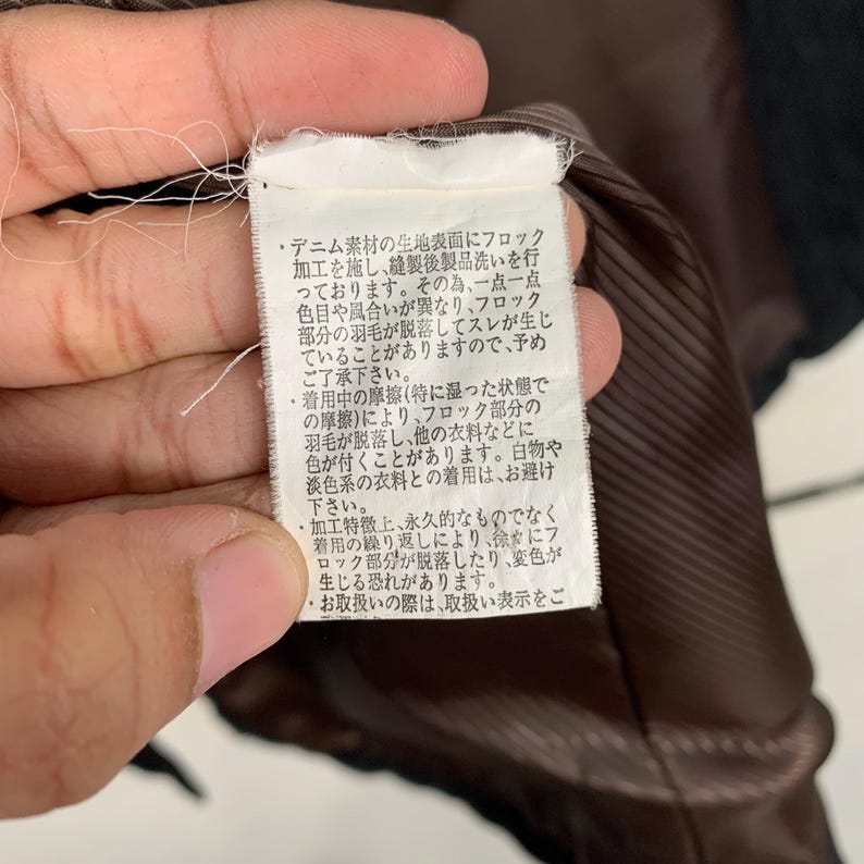May include: Close-up of a brown fabric care label with Japanese text describing the fabric's properties and care instructions. The text mentions that the fabric is denim with a flocking process, and that the color may vary due to the dyeing process. It also warns that friction may cause the flocking to come off and stain other fabrics.