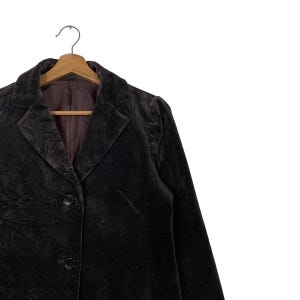 Peut inclure: Un blazer en velours marron fonc&eacute; avec un revers crant&eacute; classique et trois boutons. Le blazer est suspendu &agrave; un cintre en bois. La doublure int&eacute;rieure est d'une nuance de marron plus fonc&eacute;e.