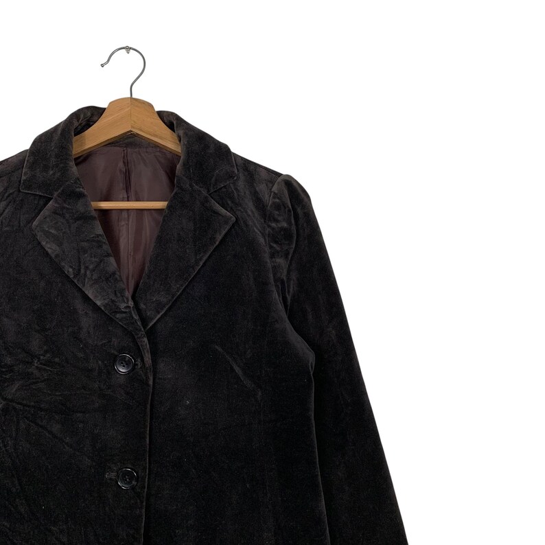 Peut inclure: Un blazer en velours marron fonc&eacute; avec un revers crant&eacute; classique et trois boutons. Le blazer est suspendu &agrave; un cintre en bois. La doublure int&eacute;rieure est d'une nuance de marron plus fonc&eacute;e.