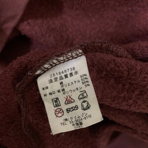 Puede incluir: Una etiqueta de tela marr&oacute;n con texto blanco e instrucciones de cuidado. La etiqueta dice "251848738 法定品質表示 本体 綿 50% ポリエステル 50%  衿・袖口・裾 綿 95% ポリウレタン 5%" e incluye varios s&iacute;mbolos para las instrucciones de lavado. La etiqueta tambi&eacute;n dice "(株)クリムゾン TEL 03-3624-9110 中国製 0182".