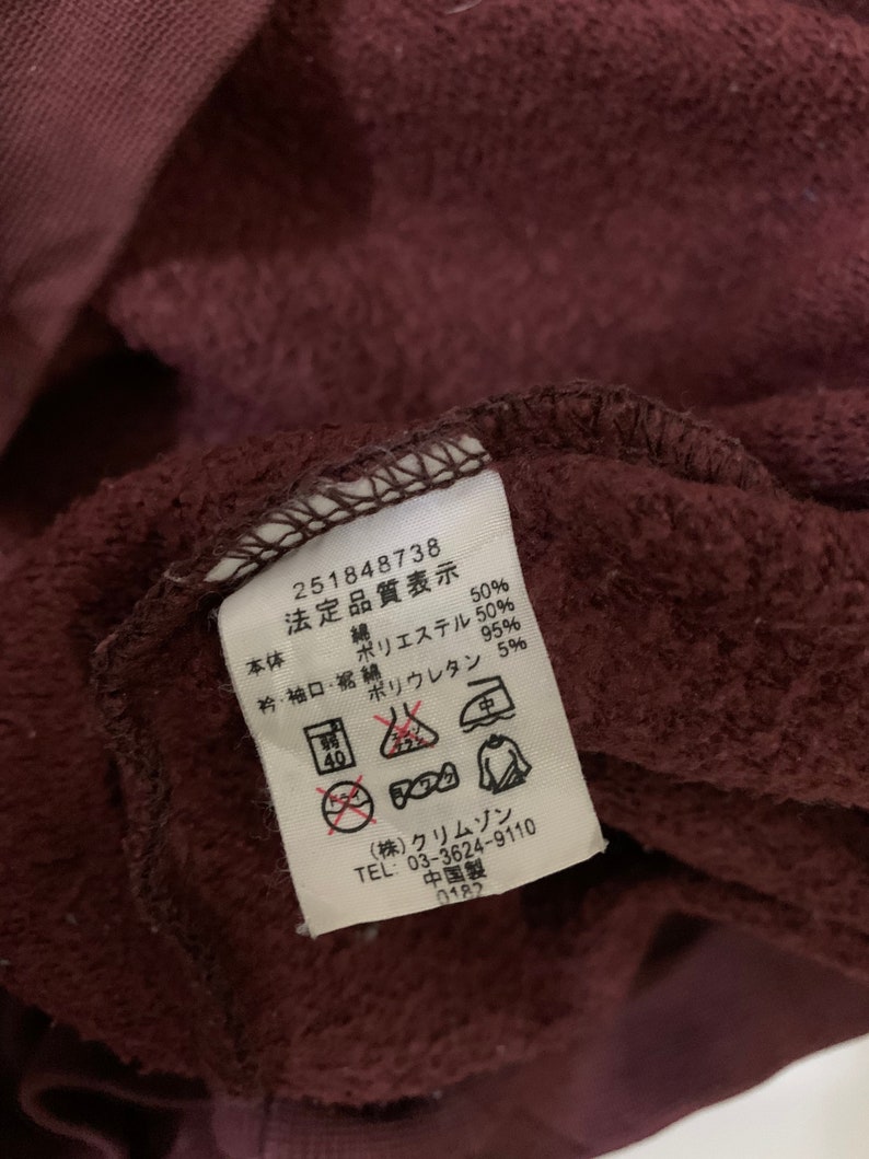 Puede incluir: Una etiqueta de tela marr&oacute;n con texto blanco e instrucciones de cuidado. La etiqueta dice "251848738 法定品質表示 本体 綿 50% ポリエステル 50%  衿・袖口・裾 綿 95% ポリウレタン 5%" e incluye varios s&iacute;mbolos para las instrucciones de lavado. La etiqueta tambi&eacute;n dice "(株)クリムゾン TEL 03-3624-9110 中国製 0182".