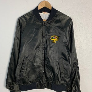 May include: A black satin bomber jacket with a black ribbed collar, cuffs, and waistband. The jacket has a snap closure and two front pockets. The jacket features an embroidered patch on the left chest that says "Indianapolis 500" with a stylized race car and checkered flag.