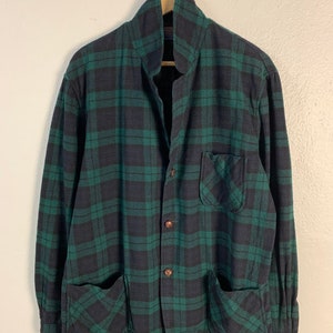 Könnte beinhalten: Ein grünes und schwarzes kariertes Flanellhemd mit Knopfleiste und zwei großen Taschen unten. Das Hemd hat einen Kragen und lange Ärmel.