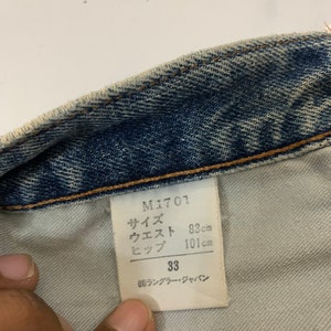Op de afbeelding: Een wit label met zwarte tekst die "M1701", "サイズ", "ウエスト", "ヒップ", "33" en "㈱ランブラー・ジャパン" luidt. Het label bevat ook metingen in centimeters: "83cm", "101cm".