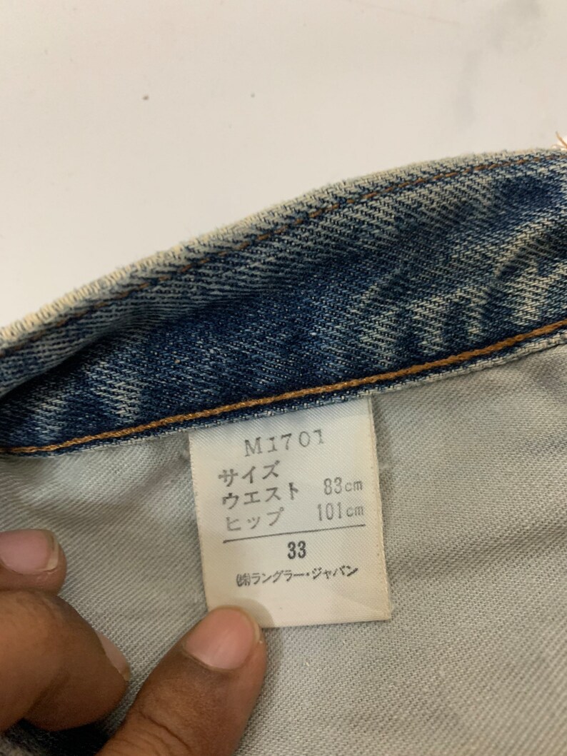 Op de afbeelding: Een wit label met zwarte tekst die "M1701", "サイズ", "ウエスト", "ヒップ", "33" en "㈱ランブラー・ジャパン" luidt. Het label bevat ook metingen in centimeters: "83cm", "101cm".