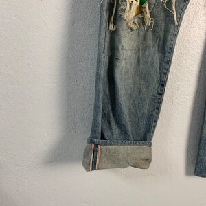 Op de afbeelding: Blauwe denim jeans met een destroyed look en een gele, groene en witte patch op het rechterbeen. De jeans hebben een gezoomde zoom met rode en witte strepen.