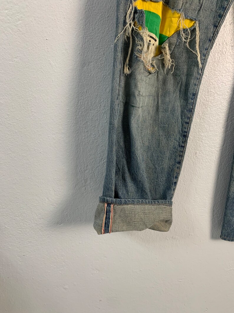 Op de afbeelding: Blauwe denim jeans met een destroyed look en een gele, groene en witte patch op het rechterbeen. De jeans hebben een gezoomde zoom met rode en witte strepen.