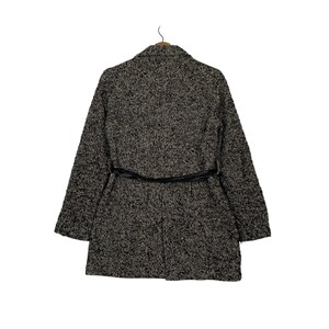 Puede incluir: Un abrigo largo texturizado en tonos negro, gris y beige, con un cintur&oacute;n negro en la cintura. El abrigo tiene un cuello cl&aacute;sico y mangas largas, perfecto para el clima fresco. El abrigo est&aacute; colgado de una percha de madera.