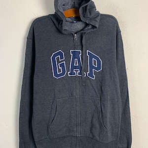 Puede incluir: Una sudadera con capucha gris oscuro con un logotipo "GAP" azul marino en la parte delantera.