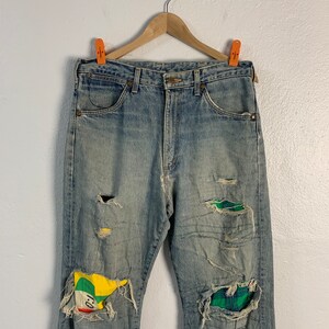 Op de afbeelding: Lichtblauwe denim jeans met meerdere opgenaaide patches. De patches zijn gemaakt van verschillende stoffen, waaronder ruitjesstof en een gele, groene en rode stof met een cartoon gezicht.
