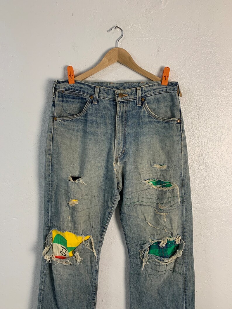 Op de afbeelding: Lichtblauwe denim jeans met meerdere opgenaaide patches. De patches zijn gemaakt van verschillende stoffen, waaronder ruitjesstof en een gele, groene en rode stof met een cartoon gezicht.