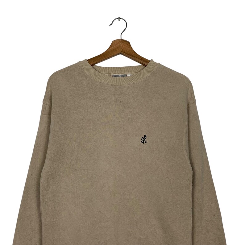 May include: A beige long-sleeved sweatshirt with a small black embroidered logo on the front.