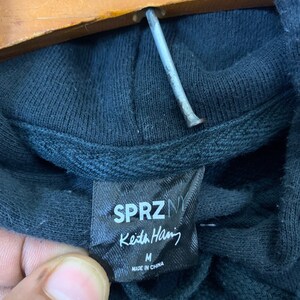 May include: A black hoodie with a SPRZNY Keith Haring tag. The tag is black with white text and says "SPRZNY Keith Haring" and "M" for size. The tag also says "Made in China".