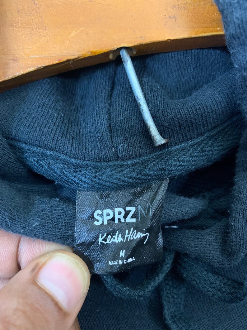 May include: A black hoodie with a SPRZNY Keith Haring tag. The tag is black with white text and says "SPRZNY Keith Haring" and "M" for size. The tag also says "Made in China".