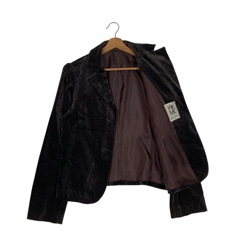 Peut inclure: Un blazer en velours marron fonc&eacute; avec un revers crant&eacute; et une fermeture &agrave; un bouton. La veste a une coupe ajust&eacute;e, des manches longues et une doublure en satin marron. Le blazer est suspendu &agrave; un cintre en bois.