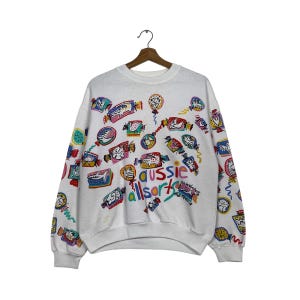 Könnte beinhalten: Weißer Pullover mit einem bunten Allover-Druck aus Süßigkeiten, Tieren und dem Text "Aussie Allsorts".