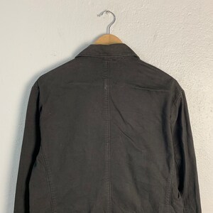Puede incluir: Un blazer negro colgado de una percha de madera. El blazer tiene un cuello cl&aacute;sico y mangas largas. La parte trasera del blazer es visible, mostrando las costuras y los detalles de la confecci&oacute;n. La tela parece ser un algod&oacute;n resistente o una mezcla.