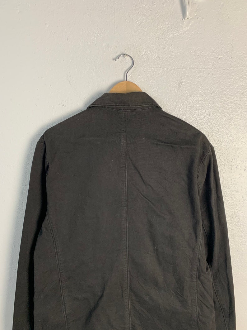 Puede incluir: Un blazer negro colgado de una percha de madera. El blazer tiene un cuello cl&aacute;sico y mangas largas. La parte trasera del blazer es visible, mostrando las costuras y los detalles de la confecci&oacute;n. La tela parece ser un algod&oacute;n resistente o una mezcla.