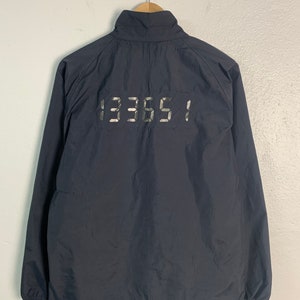 Puede incluir: Una chaqueta azul oscuro con cierre de cremallera y un gráfico reflectante plateado '133651' en la espalda.