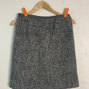 May include: A grey tweed skirt with a black waistband. The skirt is hanging on a wooden hanger.