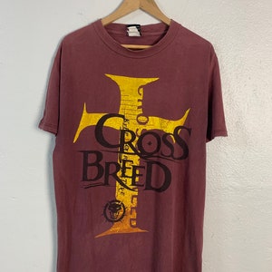 Könnte beinhalten: Ein maroonfarbenes T-Shirt mit einem goldenen Kreuz-Motiv und dem Text "Cross Breed" in Schwarz. Das Kreuz-Motiv hat einen Totenkopf am unteren Ende.