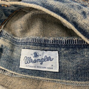 Op de afbeelding: Een close-up van een blauw denim Wrangler jeanslabel met de tekst "Wrangler Authentic Western Wear" en de nummers "19" en "64" aan weerszijden van een wereldlogo.