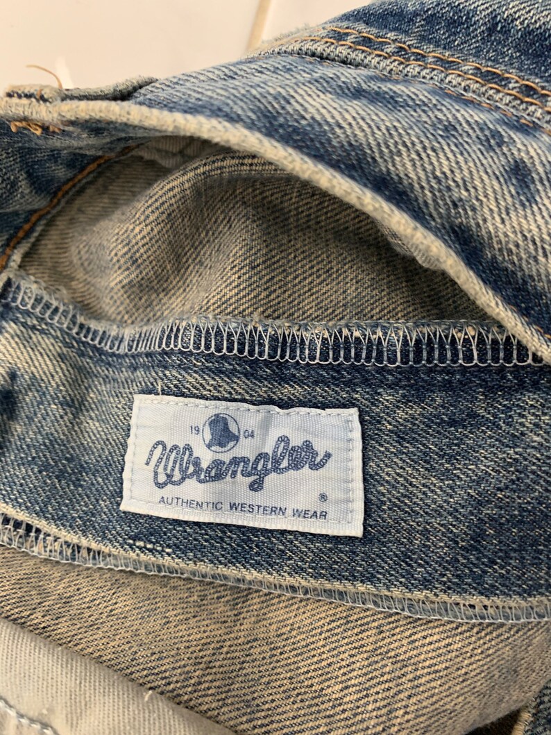 Op de afbeelding: Een close-up van een blauw denim Wrangler jeanslabel met de tekst "Wrangler Authentic Western Wear" en de nummers "19" en "64" aan weerszijden van een wereldlogo.