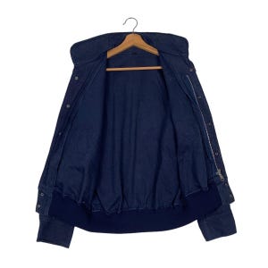 May include: A navy blue jacket with a high collar and a zipper closure. The jacket has a bomber-style bottom hem and cuffs. It is hanging on a wooden hanger against a white background. The jacket is open, revealing the interior lining.