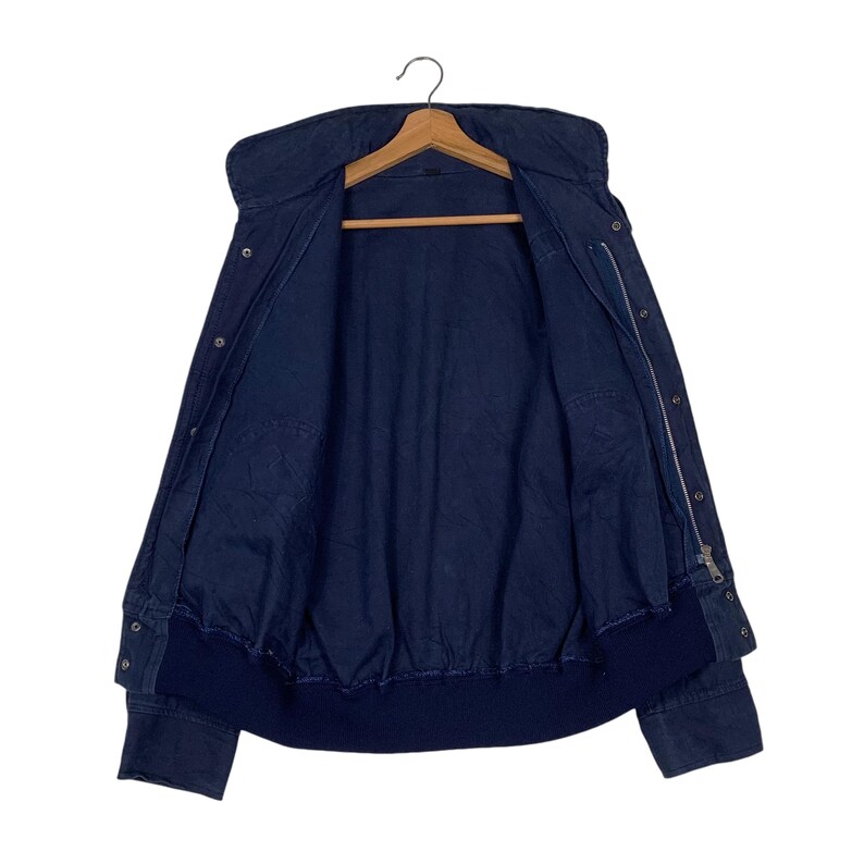 May include: A navy blue jacket with a high collar and a zipper closure. The jacket has a bomber-style bottom hem and cuffs. It is hanging on a wooden hanger against a white background. The jacket is open, revealing the interior lining.